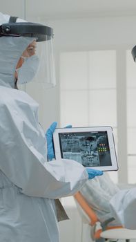 Team of dentists wearing ppe suits while looking at x ray teeth scan on digital tablet for orthodontic consultation. Caucasian stomatologists examining dental patient radiography