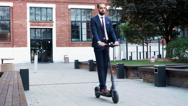 Executive manager in diplomatic suit riding on electric scooter in front of startup business company office. Entrepreneur man going to management meeting. Concept of eco-friendly transportation