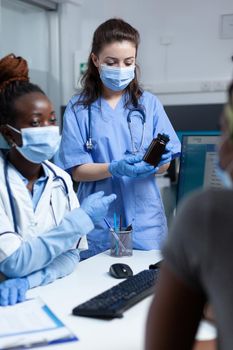 Hospital team discussing with african american sick patient explaining medication treatment during clinical consultation in medical office. Women wearing protection face mask against coronavirus