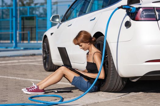 Girl waiting on the ground while her electric car is charging. Working on a laptop computer.