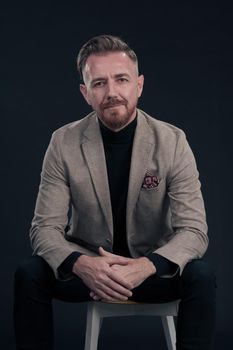 Portrait of adult businessman wearing trendy suit and sitting in modern studio on stylish chair against the black background. Horizontal mockup. High quality photo