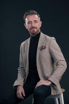 Portrait of adult businessman wearing trendy suit and sitting in modern studio on stylish chair against the black background. Horizontal mockup. High quality photo