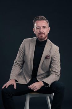 Portrait of adult businessman wearing trendy suit and sitting in modern studio on stylish chair against the black background. Horizontal mockup. High quality photo