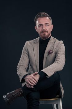 Portrait of adult businessman wearing trendy suit and sitting in modern studio on stylish chair against the black background. Horizontal mockup. High quality photo