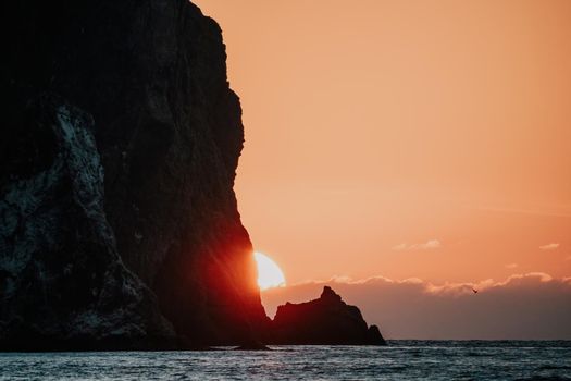 A red burning sunset with the silhouette of a cliff over the sea. Abstract nature summer or spring ocean sea background. Small waves on golden warm water surface with bokeh lights from sun.