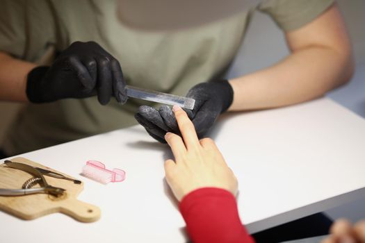 Close-up of nail master using nail file tool to create perfect shape for clients nails. Worker wear protective black gloves for work. Beauty studio concept