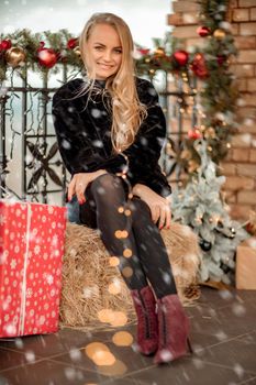 Young blonde with long hair near the cafe, decorated for Christmas. She is dressed in a black short fur coat and a blue short dress