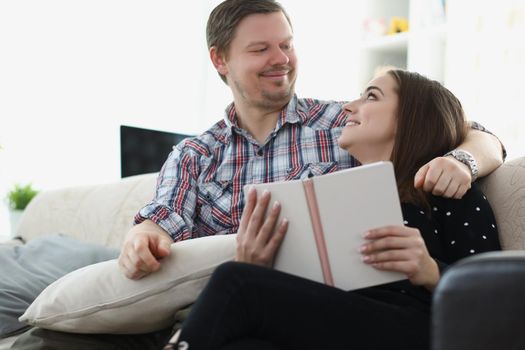 Portrait of happy couple spending time together at home in love with each other. Man kindly looking on woman, reading book on couch. Family, love concept