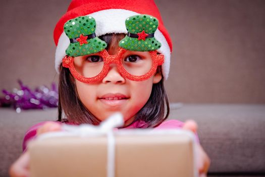 Happy little girl in Santa hat giving a Christmas present at home. Happy New Year and Merry Christmas.