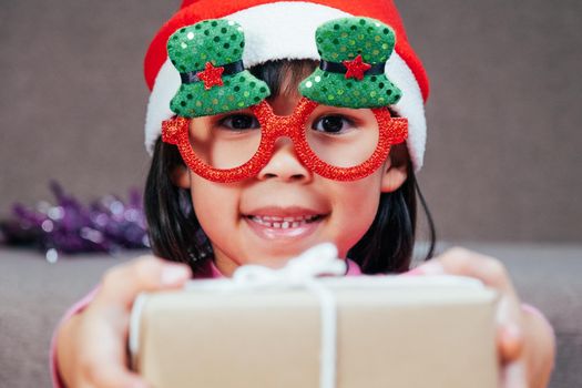 Happy little girl in Santa hat giving a Christmas present at home. Happy New Year and Merry Christmas.