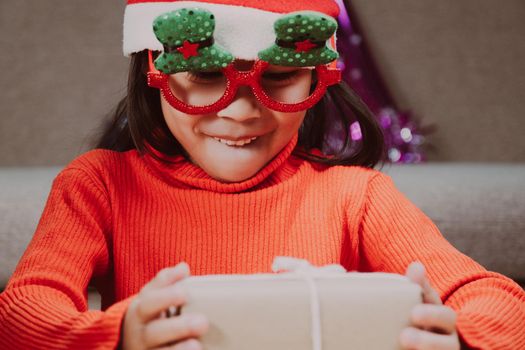 Little girl in Santa hat opening gift box sitting in the living room with Christmas tree at home. Happy New Year and Merry Christmas.