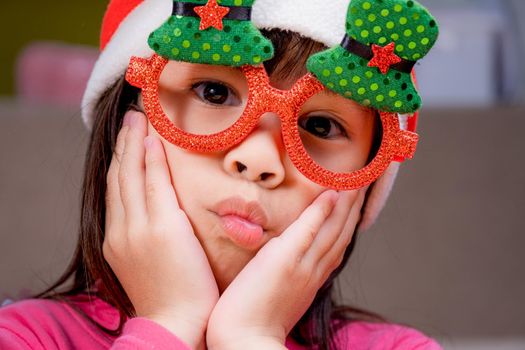 Little girl in a Santa hat sitting on the floor and making wishes near the Christmas tree at home. Happy New Year and Merry Christmas.