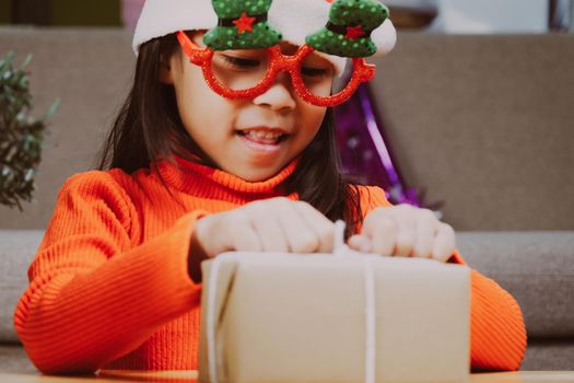 Little girl in Santa hat opening gift box sitting in the living room with Christmas tree at home. Happy New Year and Merry Christmas.