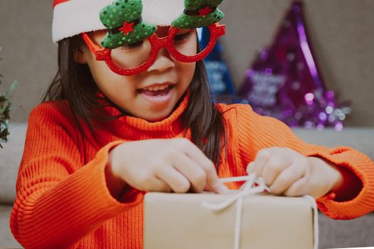 Little girl in Santa hat opening gift box sitting in the living room with Christmas tree at home. Happy New Year and Merry Christmas.