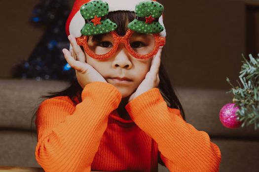 Little girl in a Santa hat sitting on the floor and making wishes near the Christmas tree at home. Happy New Year and Merry Christmas.