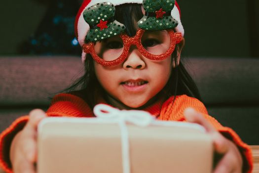 Happy little girl in Santa hat giving a Christmas present at home. Happy New Year and Merry Christmas.