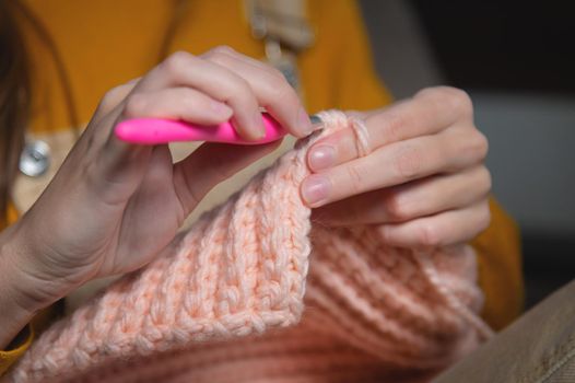 A young Caucasian woman in casual home clothes knits a pink wool hat or scarf. A useful hobby is home production of clothes. Close-up.