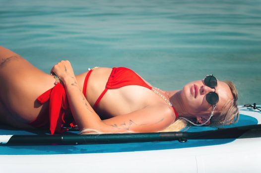 Young sexy surfer woman in red swimsuit and sunglasses lying on her sup board. Relax on stand up paddle on a sunny day Camping concept.