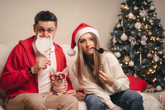 Portrait of happy family with puppy holding festive party props for photo booth. Couple woman and man in Santa hat and dog in sweater having fun on Christmas holidays at home.