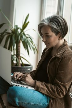 Middle aged attractive business woman working on laptop compute sitting on a windowsill at free woking zone or workstation. A beautiful aged woman runs her own business as a freelancer.