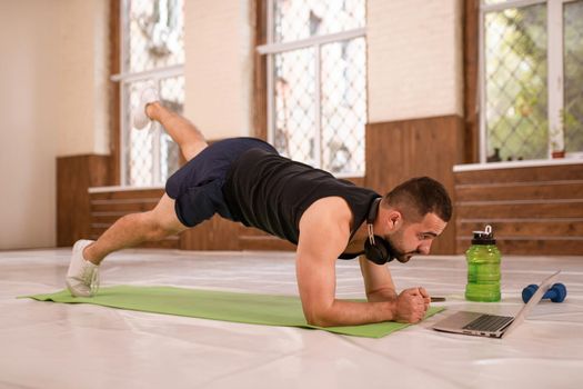 Young fitness man doing one leg up push ups exercise in empty gym or home watching online sports videos all in black clothes. Muscular sportsman doing exercises alone in gym.