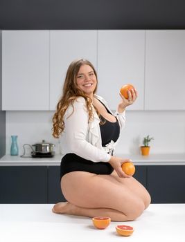 Healthy food happy curvy body young woman with long blond hair juggling fruits sitting on modern kitchen table, blending fresh fruits for healthy smoothie. Dieting and nutrition concept.