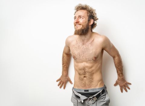 Shirtless handsome tattooed young guy with beard and curly hair wearing unbuttoned denim jeans with underwear under looking sideways isolated on white background.