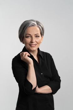 Grey haired mid aged pretty woman looking at camera with arms folded wearing black shirt isolated on grey background. Confident mature grey haired woman. Human emotions, facial expression concept.