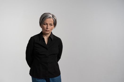 Distressed or disappointed mature grey haired woman looking down. Pretty mid aged grey haired woman in black shirt isolated on grey background. Human emotions, facial expression concept.