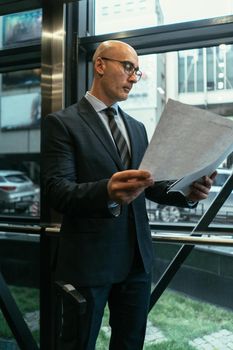 Business man looking at papers next to the window. Businessman looking at documents wearing a suit standing next to the window with suitcase next to him in the modern office with street view.