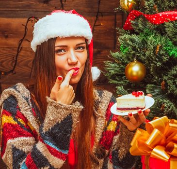 Bright lady holding a plate with a slice of cake and tasting it, finger in the mouth, Christmas Eve, dinner, cake, Tree, Christmas Gifts, Wood Background. Close-up. High quality photo