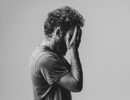 Drama, crisis, tired exhausted man cover face with his hands. Lost face young man woke up in the morning man with curly hair in olive t-shirt standing sideways isolated on white background. Black and white image.