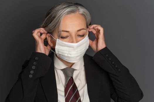 Grey-haired middle-aged business woman putting on a white medical protective mask looking down. Portrait of modern senior woman in studio wearing business clothes isolated on grey background.