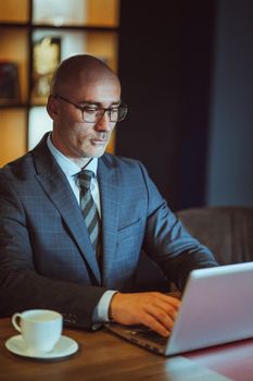 Middle aged Bald businessman in suite working on laptop sitting in the modern office. Handsome bold man working on computer in office with a cup of coffee on the table.