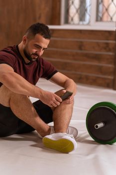 Handsome man sitting on a floor with smartphone in his hands and black and green tone fitness barbell, equipment for weight training concept. Sports equipment for training. Healthy lifestyle concept.