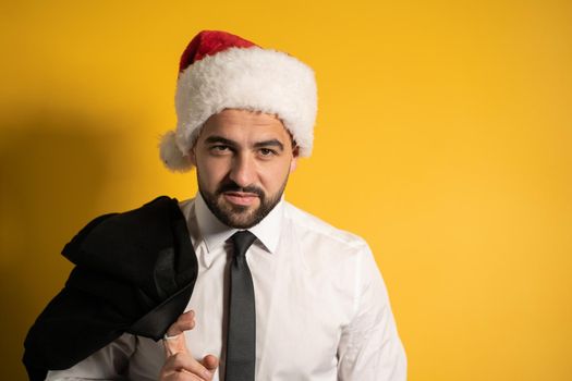 Handsome young man wearing Santa hat during Christmas season and is ready to congratulate the coworkers. Handsome smiling bearded man in Santa hat isolated on yellow background.