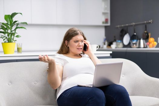 Talking on the phone while working on laptop sitting at home overweight young woman staying at home during quarantine. Self isolation. Working distantly from home using laptop.