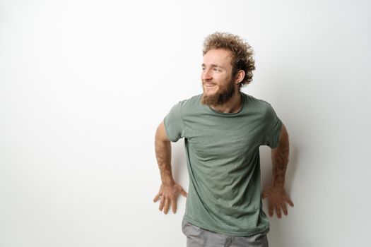 Leaned on the wall and looking sideways handsome young man with curly hair in olive t-shirt isolated on white background. Portrait of smiling young man with hands on the wall.
