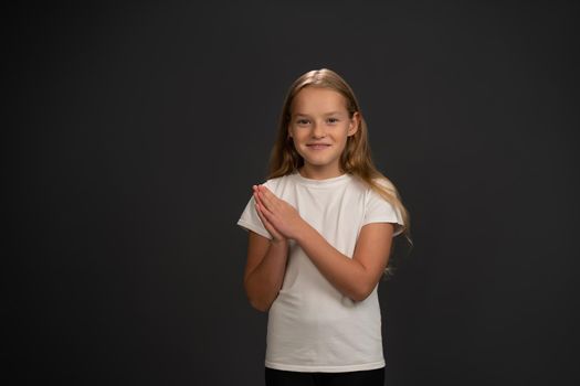 Cheering little girl standing with hands put together happily looking at the camera wearing white t-shirt isolated on dark grey or black background.
