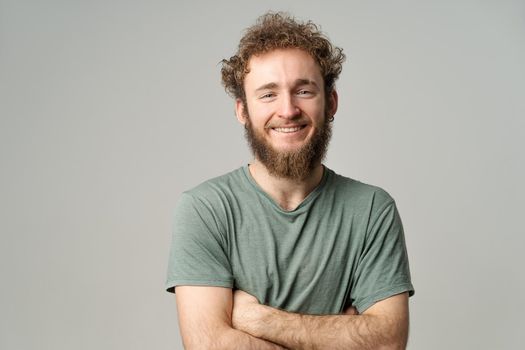 Young handsome man with beard and curly hair in olive t-shirt looking at camera isolated on white background. Portrait of smiling young man with hands folded.