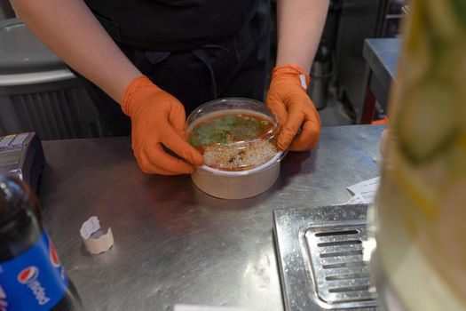 Worker wearing gloves packs tom yum soup into container. Asian restaurant. June 2020. Kyiv, Ukraine.