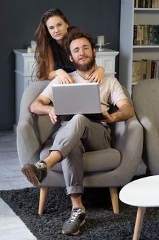 Working together young couple staying quarantined at home with sitting on the arm chairs. Happy young couple sitting on a chair together using laptop. Square cropped.