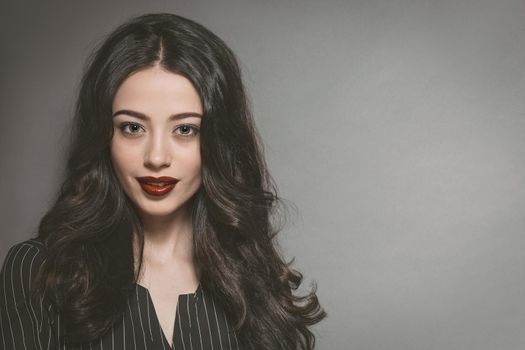 Pretty business woman in black white stripes jacket and beautiful dark hair isolated on grey background. Young female model with professional make-up. Cosmetics.