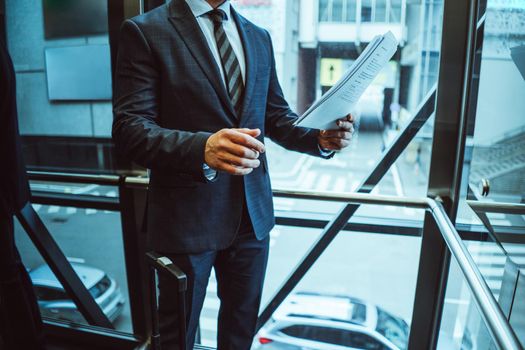 Looking thru documents businessman in suit standing next to the window with suitcase next to him in the modern office with street view. Business man looking at papers next to the window.