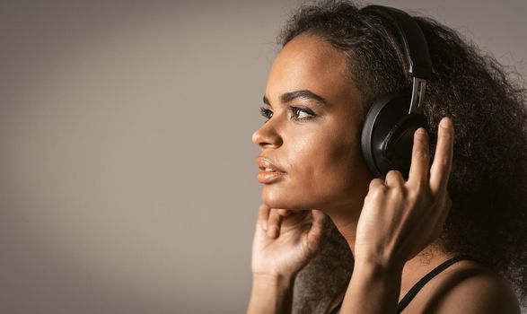 Charming young African-American girl listening her favourite track in headphones wearing red jacket black top under isolated on grey background, emotionally move, have fun. Cold toned image.