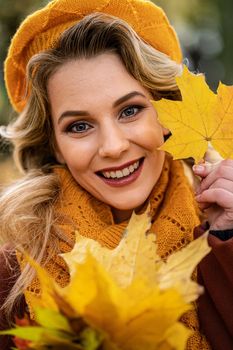 Close up. Young woman in beige knitted beret with autumn leaves in hand and fall yellow garden or park. Beautiful smiling young woman in autumn leaves.