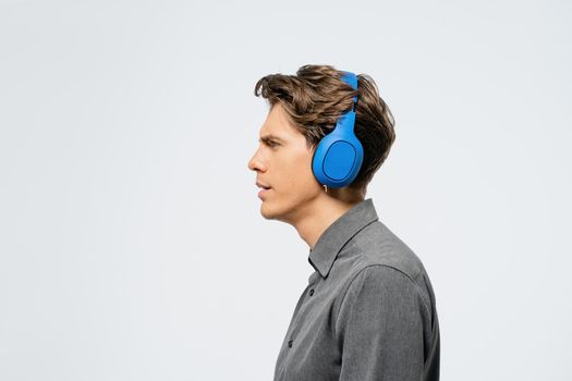 Portrait of a young guy in grey outfit standing sideways listening music wearing blue wireless headphones. Funny young guy listen to his favourite track or song.