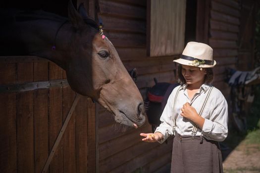 Charming young girl in vintage clothes standing with her brown horse sticking out head of the barn at sunny daytime. Friendship concept.