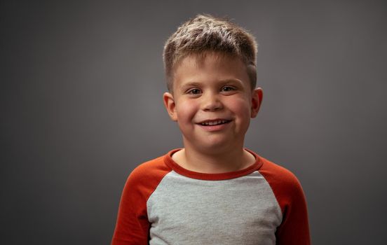 Pretty preschooler boy in red shirt smiling on camera. Happy kid on dark grey background. Funny family time. Little boy enjoying having fun in studio.