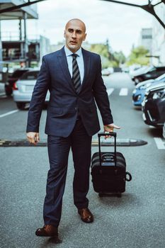 Businessman in a suit on a parking lot carrying suitcase walking thru parked cars. Businessman in suit and suitcase on business trip. Man walking outdoors with luggage determined and confident.